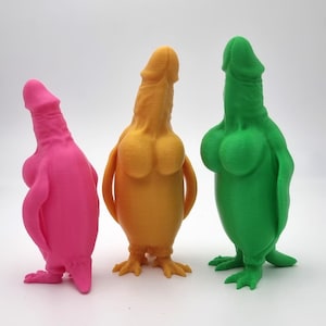 Può includere: Tre sculture dai colori vivaci in rosa, giallo e verde. Ogni scultura ha una forma fallica con un corpo e piedi simili a quelli di un uccello. Le sculture sono su una superficie bianca.