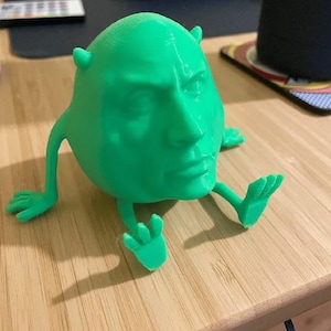 Op de afbeelding: Een groene 3D-geprinte figuur met een gezicht dat lijkt op een beroemdheid. De figuur heeft kleine hoorns, armen en voeten. Het rust op een houten oppervlak.
