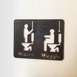 May include: Black and white bathroom sign with the words "Wizard" and "Muggle" below stick figure illustrations. The sign is rectangular with rounded corners and is mounted on a white surface.