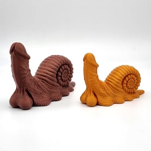 Può includere: Due sculture originali a forma di lumaca. Una è marrone scuro, l'altra arancione. I corpi delle lumache sono fallici, con una superficie strutturata e un guscio a spirale.