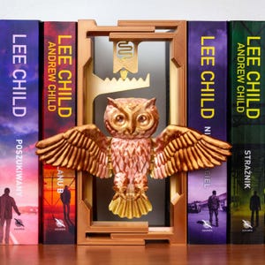 May include: A decorative owl bookend with outstretched wings, in shades of gold and copper, stands between several books. The books have titles by Lee Child and Andrew Child. The bookend is designed to hold books upright.