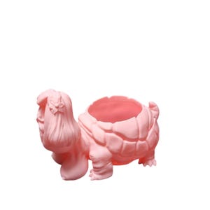 May include: A light pink turtle-shaped planter with a detailed shell and a head sculpted to resemble a woman with long hair. The planter has a large opening for plants and is made of a smooth material.