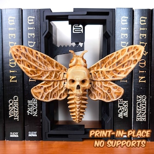 Pode incluir: Um suporte de livros decorativo em forma de traça castanha clara com um design de caveira, colocado contra uma moldura de suporte de livros preta. As asas têm padrões detalhados. O texto "PRINT-IN-PLACE NO SUPPORTS" é visível.