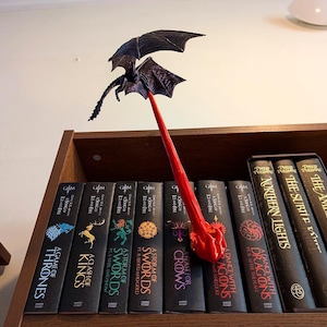 Può includere: Un fermalibri a forma di drago stampato in 3D con uno stelo rosso e un drago nero. Il fermalibri è posizionato su uno scaffale con una collezione di libri, tra cui titoli come "A Game of Thrones" e "A Clash of Kings".