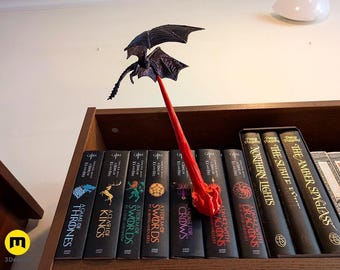 Pode incluir: Um suporte de livros de dragão impresso em 3D com uma haste vermelha e um dragão preto. O suporte de livros está colocado numa prateleira com uma coleção de livros, incluindo títulos como "A Guerra dos Tronos" e "A Fúria dos Reis".