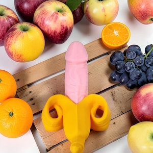 Pode incluir: Um item de novidade rosa e amarelo em forma de pênis e banana, rodeado por frutas frescas, incluindo maçãs, laranjas e uvas, dispostas sobre uma superfície de madeira.
