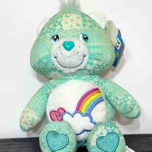 May include: A mint green Care Bear plush toy with a patchwork design. The bear has a white belly with a rainbow and cloud design, and heart-shaped accents on its feet. It has a turquoise heart on its nose and a tuft of hair on its head.