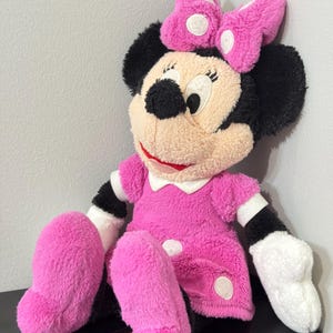 Peut inclure: Une poup&eacute;e en peluche Minnie Mouse, v&ecirc;tue d'une robe rose &agrave; pois blancs et d'un n&oelig;ud assorti. La poup&eacute;e a des oreilles noires, un visage beige et des gants blancs. La poup&eacute;e mesure environ 46 cm de haut.