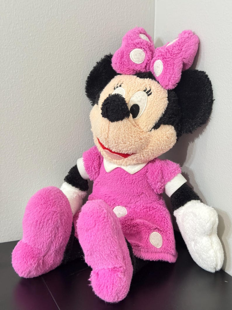 Peut inclure: Une poup&eacute;e en peluche Minnie Mouse, v&ecirc;tue d'une robe rose &agrave; pois blancs et d'un n&oelig;ud assorti. La poup&eacute;e a des oreilles noires, un visage beige et des gants blancs. La poup&eacute;e mesure environ 46 cm de haut.