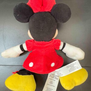 Pu&ograve; includere: Una bambola di peluche di Minnie Mouse, con un vestito rosso a pois bianchi, un fiocco rosso e orecchie nere. La bambola ha guanti bianchi e scarpe gialle. Un'etichetta con testo &egrave; attaccata alla bambola.