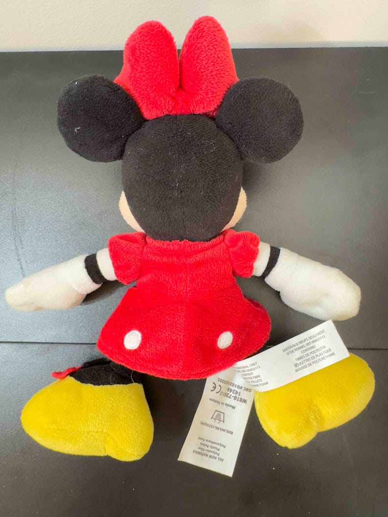 Pu&ograve; includere: Una bambola di peluche di Minnie Mouse, con un vestito rosso a pois bianchi, un fiocco rosso e orecchie nere. La bambola ha guanti bianchi e scarpe gialle. Un'etichetta con testo &egrave; attaccata alla bambola.
