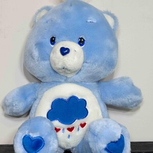 May include: A light blue plush Care Bear with a white belly featuring a blue cloud with red heart raindrops. The bear has a blue heart on its paws and nose. It has blue eyes and a tuft of blue hair.