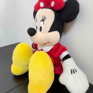 Pu&ograve; includere: Una bambola di peluche di Minnie Mouse, con un vestito rosso, un colletto bianco e un fiocco rosso a pois bianchi. La bambola ha orecchie nere, un naso nero e guanti bianchi con dettagli neri. Sono visibili i piedi gialli della bambola.