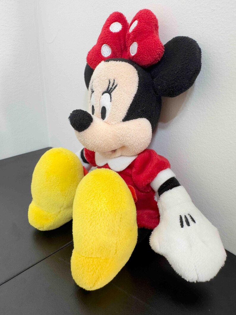Pu&ograve; includere: Una bambola di peluche di Minnie Mouse, con un vestito rosso, un colletto bianco e un fiocco rosso a pois bianchi. La bambola ha orecchie nere, un naso nero e guanti bianchi con dettagli neri. Sono visibili i piedi gialli della bambola.