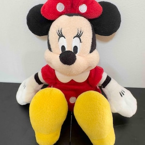Pu&ograve; includere: Un pupazzo di peluche di Minnie Mouse. La bambola ha un vestito rosso con colletto bianco, orecchie nere e un fiocco rosso a pois bianchi. I piedi della bambola sono gialli e le braccia e il viso sono color crema.