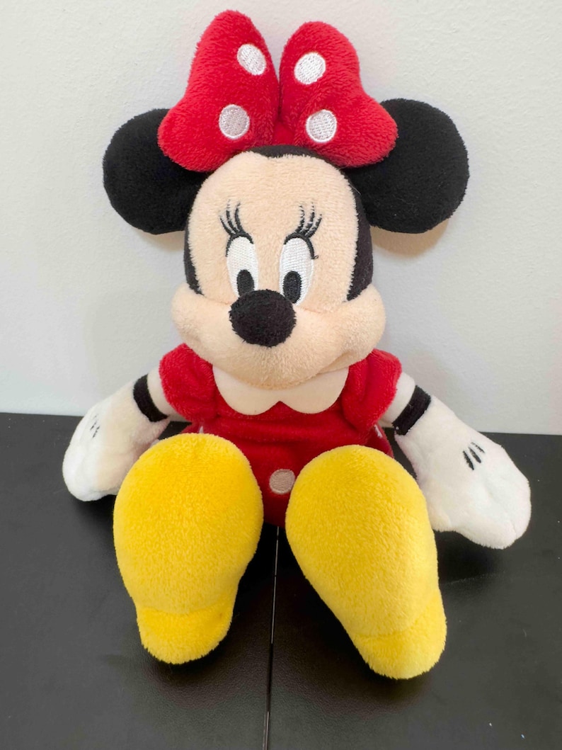 Pu&ograve; includere: Un pupazzo di peluche di Minnie Mouse. La bambola ha un vestito rosso con colletto bianco, orecchie nere e un fiocco rosso a pois bianchi. I piedi della bambola sono gialli e le braccia e il viso sono color crema.