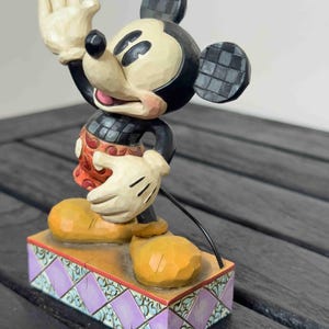 Puede incluir: Una figura de Mickey Mouse con la mano levantada, vistiendo un atuendo rojo y negro, zapatos amarillos y guantes blancos. La figura se encuentra sobre una base decorativa con detalles morados y turquesas. Las orejas tienen un patr&oacute;n de cuadros.