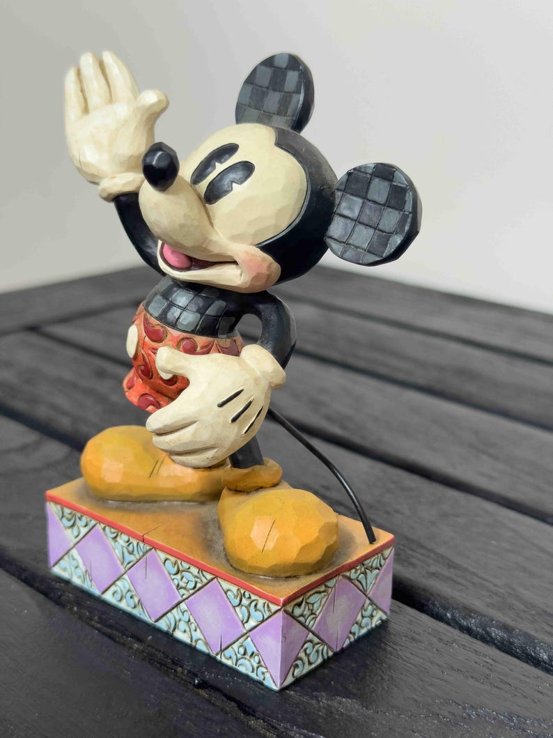 Puede incluir: Una figura de Mickey Mouse con la mano levantada, vistiendo un atuendo rojo y negro, zapatos amarillos y guantes blancos. La figura se encuentra sobre una base decorativa con detalles morados y turquesas. Las orejas tienen un patr&oacute;n de cuadros.