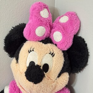 Peut inclure: Poup&eacute;e en peluche Minnie Mouse avec un n&oelig;ud rose &agrave; pois blancs. La poup&eacute;e a des oreilles noires, un visage beige et une robe rose. Les yeux de la poup&eacute;e sont grands et noirs, et elle a un col blanc.