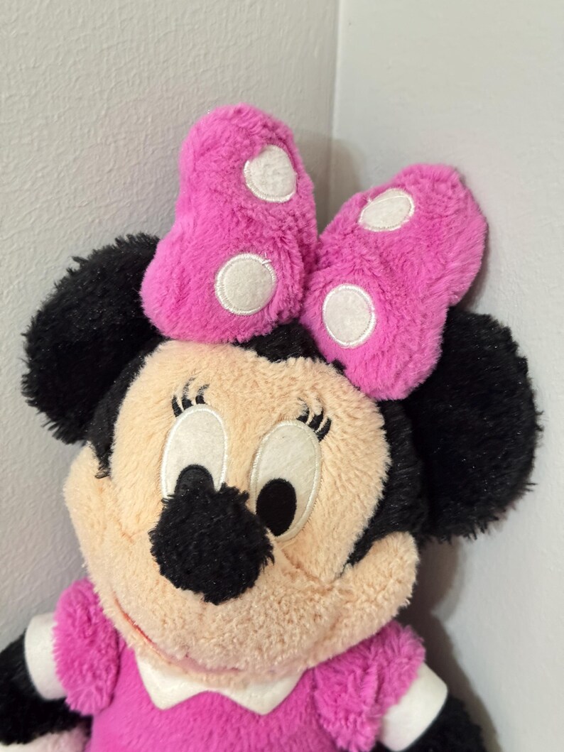 Peut inclure: Poup&eacute;e en peluche Minnie Mouse avec un n&oelig;ud rose &agrave; pois blancs. La poup&eacute;e a des oreilles noires, un visage beige et une robe rose. Les yeux de la poup&eacute;e sont grands et noirs, et elle a un col blanc.