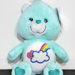May include: A light blue Care Bear plush toy with a white belly featuring a rainbow and cloud design. The bear has a heart on each foot and a heart-shaped nose. It has a smiling face and a small tuft of hair on its head.