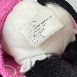 Peut inclure: Gros plan sur un jouet en peluche avec un centre blanc, des accents noirs et une bordure rose. Une &eacute;tiquette avec du texte est visible, indiquant les mat&eacute;riaux et les instructions d'entretien. Le jouet semble &ecirc;tre fait de mat&eacute;riaux doux et moelleux.