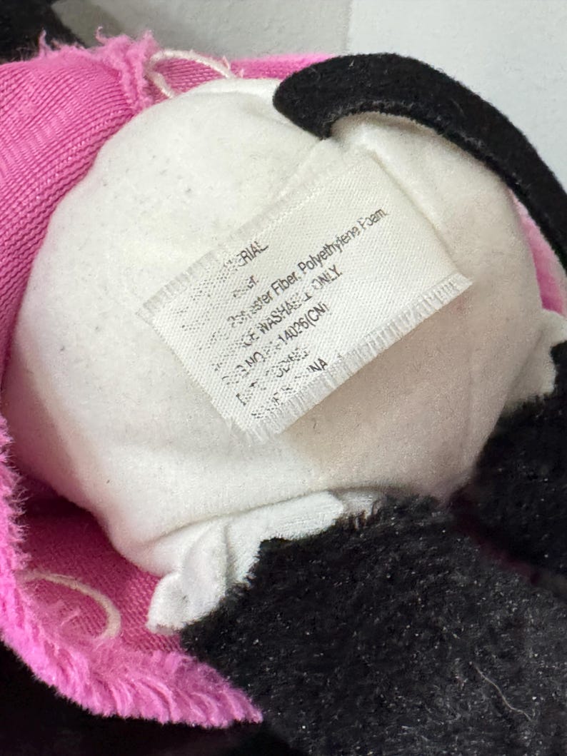 Peut inclure: Gros plan sur un jouet en peluche avec un centre blanc, des accents noirs et une bordure rose. Une &eacute;tiquette avec du texte est visible, indiquant les mat&eacute;riaux et les instructions d'entretien. Le jouet semble &ecirc;tre fait de mat&eacute;riaux doux et moelleux.