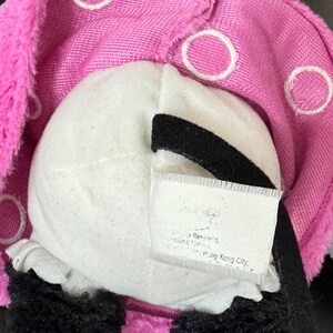 Peut inclure: Gros plan sur un jouet en peluche avec un visage blanc, des accents noirs et un couvre-chef rose &agrave; pois blancs. Le jouet a de la fourrure noire et une &eacute;tiquette avec du texte.