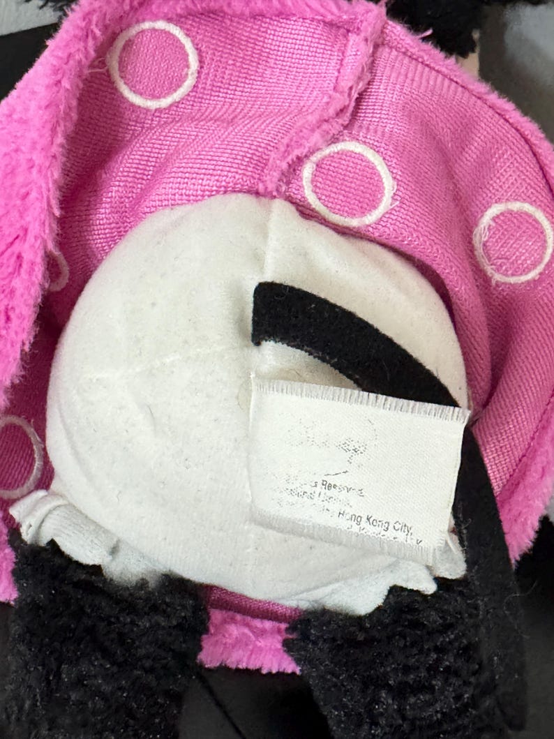 Peut inclure: Gros plan sur un jouet en peluche avec un visage blanc, des accents noirs et un couvre-chef rose &agrave; pois blancs. Le jouet a de la fourrure noire et une &eacute;tiquette avec du texte.