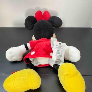 Pu&ograve; includere: Un peluche di Minnie Mouse, vista da dietro, con un vestito rosso a pois bianchi, orecchie nere e un fiocco rosso. Il giocattolo ha guanti bianchi e scarpe gialle. Un'etichetta &egrave; attaccata sul retro del peluche.