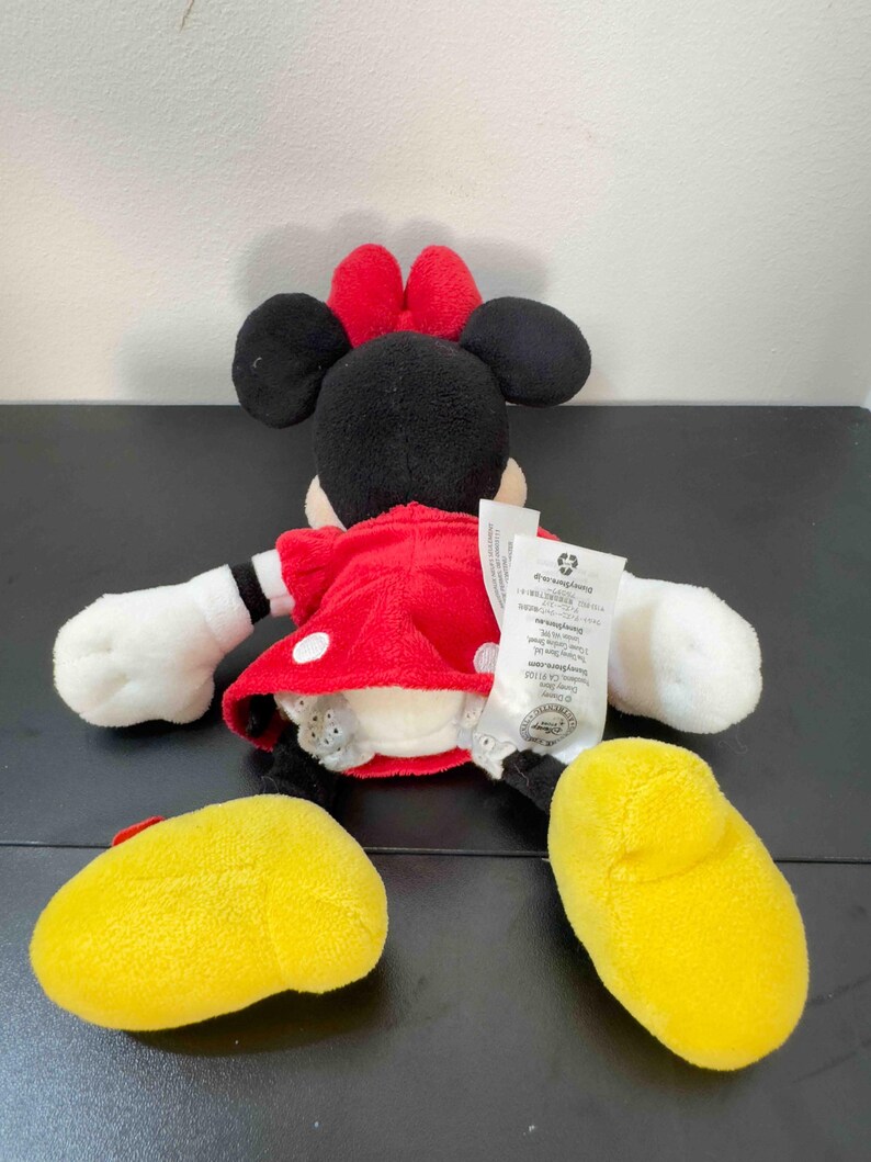 Pu&ograve; includere: Un peluche di Minnie Mouse, vista da dietro, con un vestito rosso a pois bianchi, orecchie nere e un fiocco rosso. Il giocattolo ha guanti bianchi e scarpe gialle. Un'etichetta &egrave; attaccata sul retro del peluche.