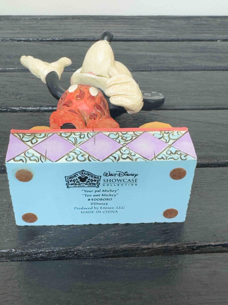 Puede incluir: Una figura de Disney Traditions con Mickey Mouse. La figura muestra a Mickey sentado, con una cabeza roja y blanca con lunares y un sombrero color crema. La base es azul claro con detalles decorativos y texto.