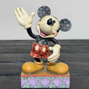 Puede incluir: Una figura de Mickey Mouse con un gesto de bienvenida. La figura tiene un cuerpo negro, guantes blancos y zapatos amarillos. Sus pantalones cortos rojos tienen lunares blancos. La figura se encuentra sobre una base decorativa con un dise&ntilde;o morado y turquesa.
