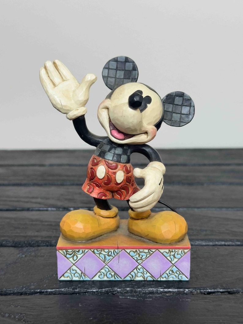 Puede incluir: Una figura de Mickey Mouse con un gesto de bienvenida. La figura tiene un cuerpo negro, guantes blancos y zapatos amarillos. Sus pantalones cortos rojos tienen lunares blancos. La figura se encuentra sobre una base decorativa con un dise&ntilde;o morado y turquesa.