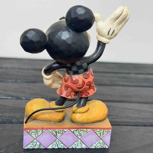 Puede incluir: Una figura de Mickey Mouse con cabeza y orejas negras, que lleva pantalones cortos rojos y un guante blanco levantado en se&ntilde;al de saludo. La figura se encuentra sobre una base decorativa con un patr&oacute;n p&uacute;rpura y verde. Los zapatos son amarillos.