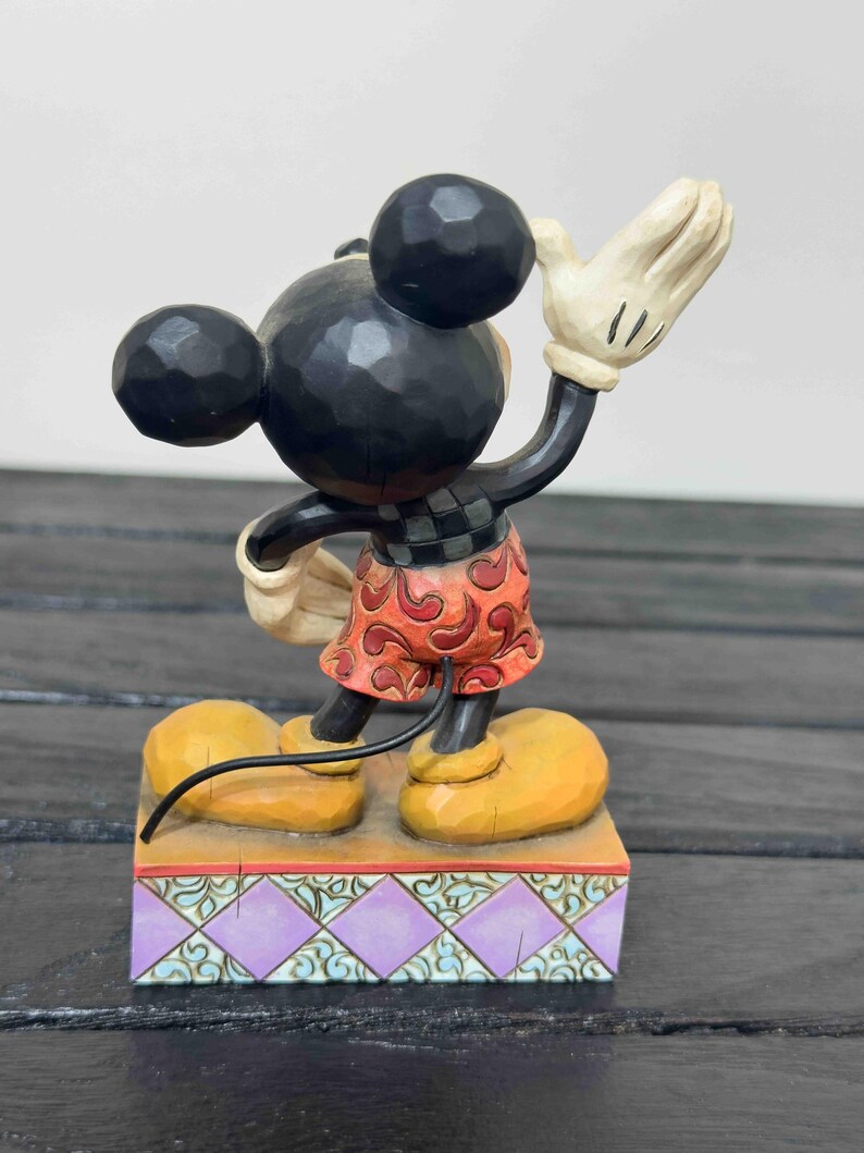 Puede incluir: Una figura de Mickey Mouse con cabeza y orejas negras, que lleva pantalones cortos rojos y un guante blanco levantado en se&ntilde;al de saludo. La figura se encuentra sobre una base decorativa con un patr&oacute;n p&uacute;rpura y verde. Los zapatos son amarillos.