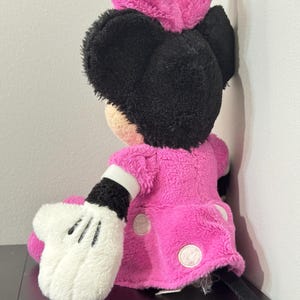 Peut inclure: Une poup&eacute;e en peluche Minnie Mouse, vue de dos, avec une robe rose vif &agrave; pois blancs et un n&oelig;ud assorti. La poup&eacute;e a des oreilles noires et une queue noire. La main gant&eacute;e blanche est visible.
