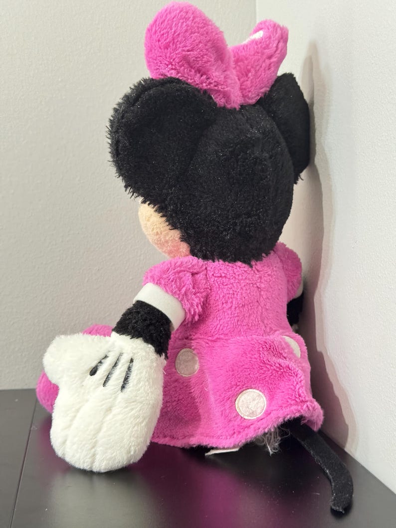 Peut inclure: Une poup&eacute;e en peluche Minnie Mouse, vue de dos, avec une robe rose vif &agrave; pois blancs et un n&oelig;ud assorti. La poup&eacute;e a des oreilles noires et une queue noire. La main gant&eacute;e blanche est visible.