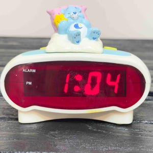 May include: A white and blue digital alarm clock with a red display showing 1:04 PM. A blue Care Bear figure rests on a cloud on top of the clock. The clock has a black power cord.