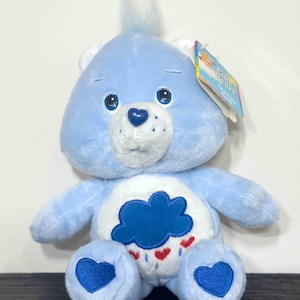 May include: A light blue Care Bear plush toy with a cloud and heart design on its belly. The bear has a blue nose, blue heart-shaped feet, and a tuft of blue hair. A tag is attached to the bear's ear.