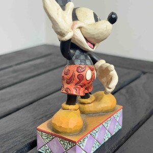 Puede incluir: Una figura de Mickey Mouse con la mano levantada, vistiendo una camisa a cuadros negros y grises, pantalones cortos rojos con lunares blancos y zapatos amarillos. La figura se encuentra sobre una base decorativa con un patr&oacute;n de diamantes morados y turquesas.