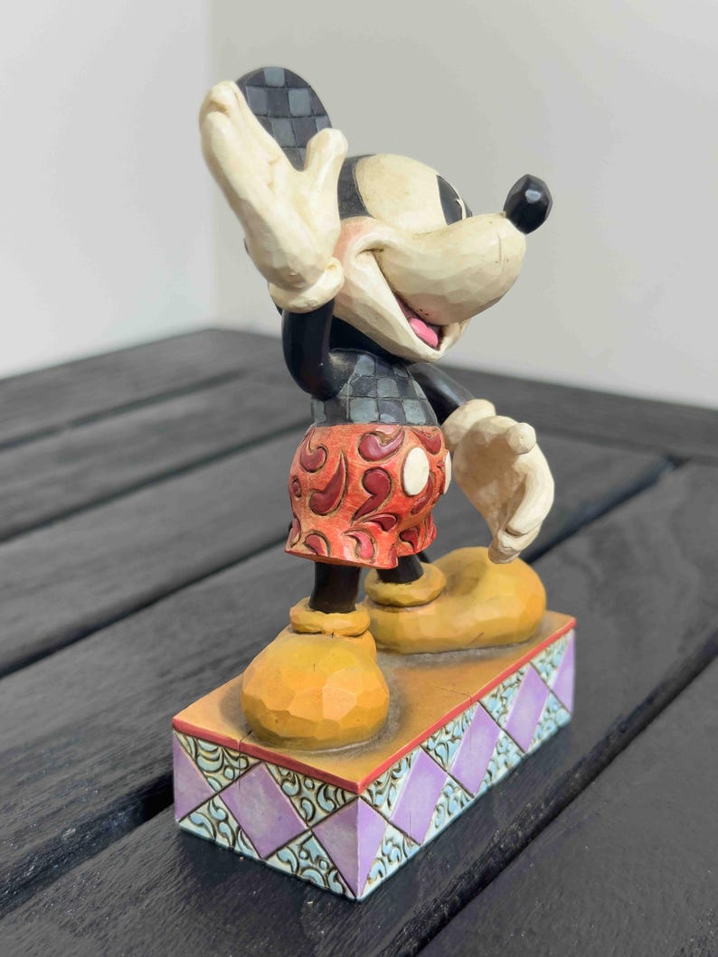 Puede incluir: Una figura de Mickey Mouse con la mano levantada, vistiendo una camisa a cuadros negros y grises, pantalones cortos rojos con lunares blancos y zapatos amarillos. La figura se encuentra sobre una base decorativa con un patr&oacute;n de diamantes morados y turquesas.