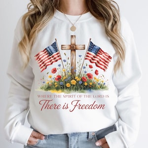 Puede incluir: Sudadera blanca con un gráfico de dos banderas americanas que flanquean una cruz de madera con un arreglo floral en la base. El texto "Where the spirit of the Lord is There is Freedom" está impreso debajo del gráfico.