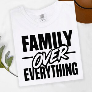 May include: A white t-shirt with the phrase "FAMILY OVER EVERYTHING" in bold black lettering. The word "OVER" is in a stylized, cursive font. A brown hat is partially visible in the background.