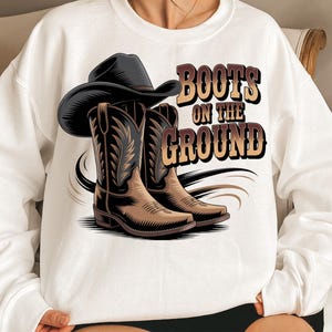 Op de afbeelding: Witte sweatshirt met een afbeelding van een zwarte cowboyhoed en bruine cowboylaarzen. De tekst "Boots on the Ground" is in bruin en beige. Het ontwerp heeft een country western thema.