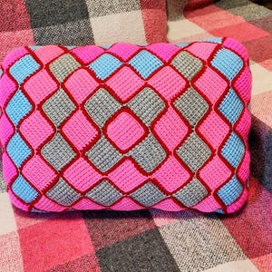 May include: A rectangular cushion with a geometric pattern. The design features a repeating diamond pattern in shades of pink, grey, and light blue, outlined with red stitching. The cushion is a decorative item.