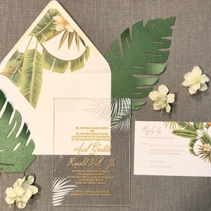 May include: A clear acrylic wedding invitation with gold lettering. The invitation features a tropical leaf design and the text "MR. AND MRS. CALVIN CORBETT AND MR. AND MRS. HILTON GLOVER REQUEST THE HONOUR OF YOUR PRESENCE AT THE MARRIAGE OF THEIR CHILDREN ARIEL CORBETT AND RONALD HILL JR. ON SUNDAY THE EIGHTH OF MARCH TWO THOUSAND TWENTY AT HALF PAST FIVE O'CLOCK IN THE EVENING THE FIVES AZUL RESORT PLAYA DEL CARMEN, MEXICO COCKTAILS, DINNER AND DANCING IMMEDIATELY FOLLOWING CEREMONY".