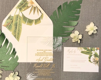 May include: A clear acrylic wedding invitation with gold lettering. The invitation features a tropical leaf design and the text "MR. AND MRS. CALVIN CORBETT AND MR. AND MRS. HILTON GLOVER REQUEST THE HONOUR OF YOUR PRESENCE AT THE MARRIAGE OF THEIR CHILDREN ARIEL CORBETT AND RONALD HILL JR. ON SUNDAY THE EIGHTH OF MARCH TWO THOUSAND TWENTY AT HALF PAST FIVE O'CLOCK IN THE EVENING THE FIVES AZUL RESORT PLAYA DEL CARMEN, MEXICO COCKTAILS, DINNER AND DANCING IMMEDIATELY FOLLOWING CEREMONY".