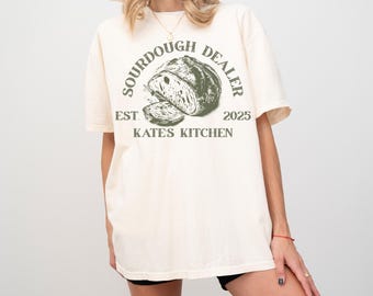 May include: A cream-coloured t-shirt featuring a green graphic of a sourdough loaf and the text "SOURDOUGH DEALER" in a circular arrangement. Beneath the bread image are the words "EST. 2025" and "KATES KITCHEN". The shirt has short sleeves and a loose fit.
