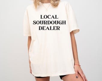 May include: A cream-coloured t-shirt with the words "LOCAL SOURDOUGH DEALER" printed in black. The shirt has short sleeves and a relaxed fit. The model is wearing black shorts.